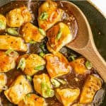 A wooden spoon resting in a large skillet with cooked cubed pieces of teriyaki chicken and a text overlay on the photo