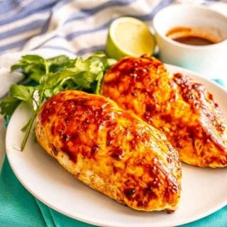 Two grilled adobo chicken breasts on a white plate with parsley, lime and a small bowl of adobo sauce nearby