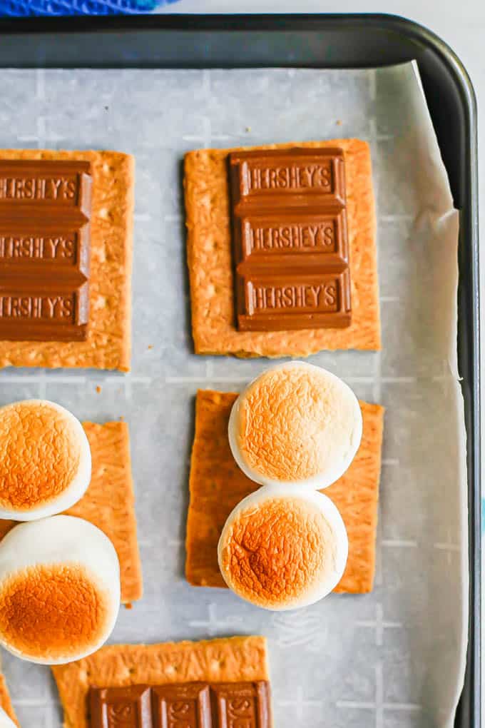 Toasted marshmallows and softened chocolate on graham crackers on a baking sheet after being baked in the oven