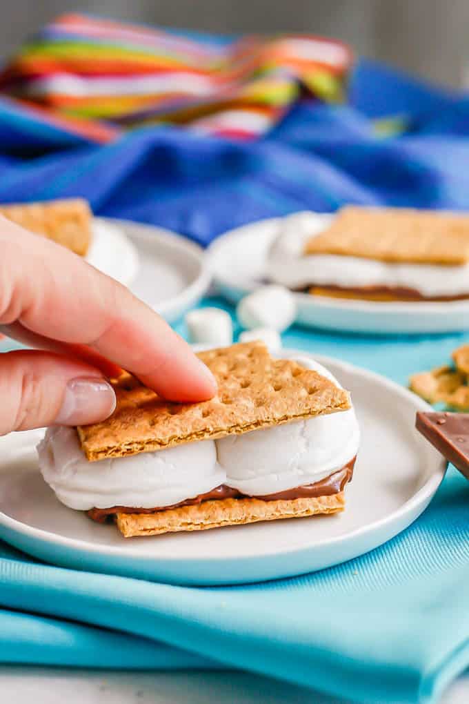 A hand smushing down a s'mores sandwich on a small white plate