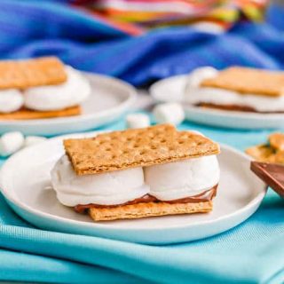 S'mores on small white plates with chocolate pieces, graham cracker pieces and mini marshmallows nearby