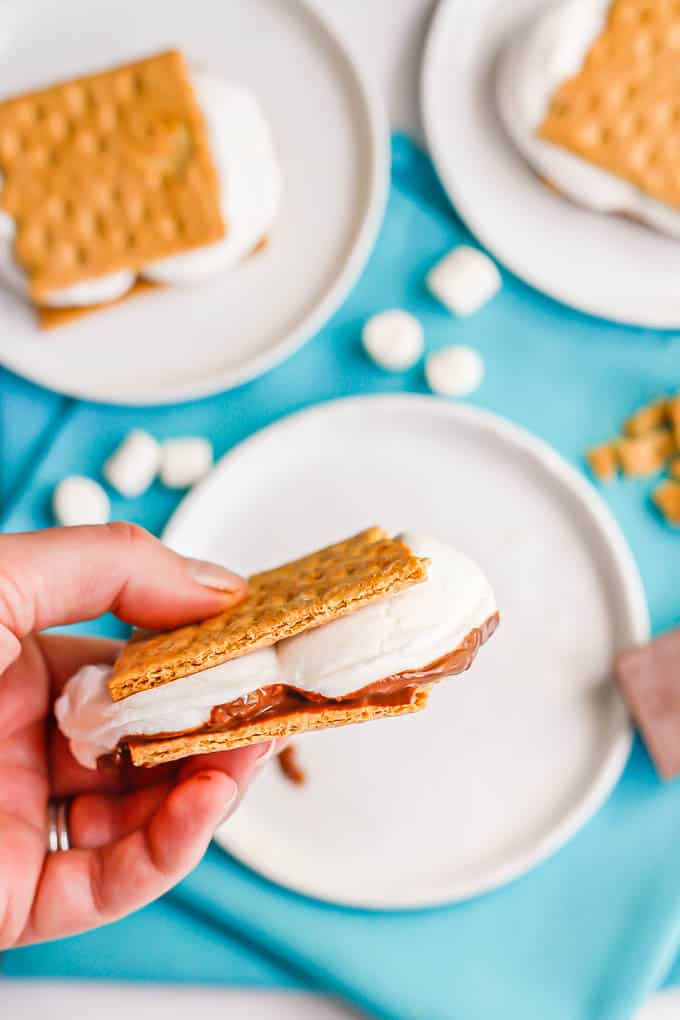 A hand picking up a melty s'mores from a plate with s'mores fixings and extra s'mores sandwiches in the background