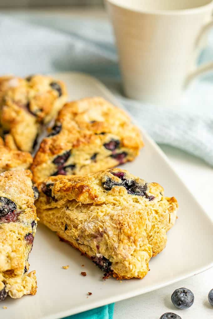 A white serving plate with triangular blueberry scones with a coffee mug in the background