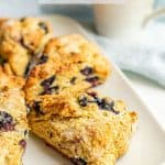 A white serving plate with triangular blueberry scones with a coffee mug in the background and a text overlay on the photo