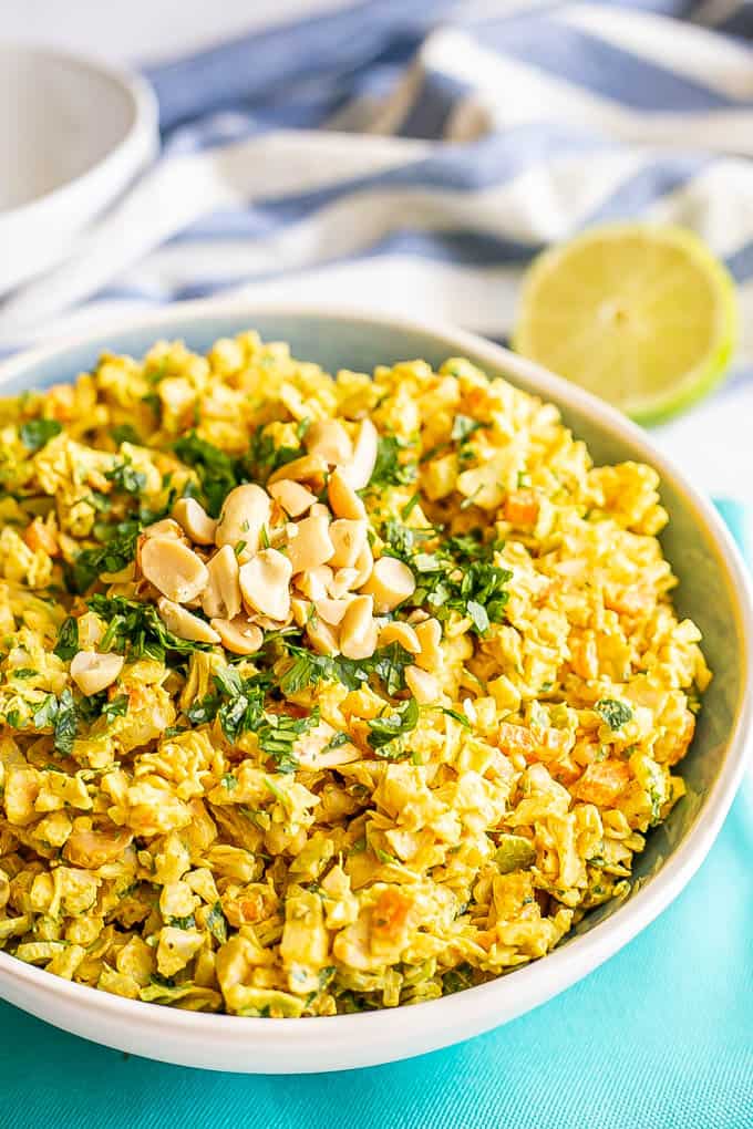 Golden curried coleslaw in a blue and white serving bowl with peanuts and cilantro on top and a halved lime in the background