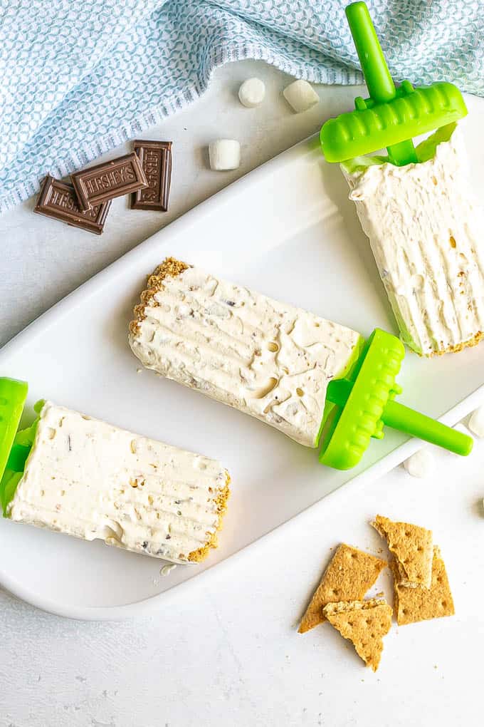 Three s'mores popsicles laid out on a white platter with graham crackers, mini marshmallows and chocolate pieces nearby