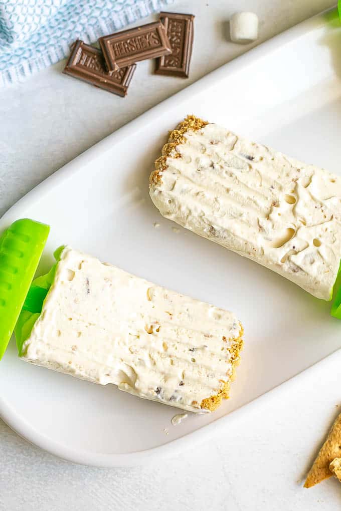 S'mores popsicles laid out on a white platter with graham crackers, mini marshmallows and chocolate pieces nearby