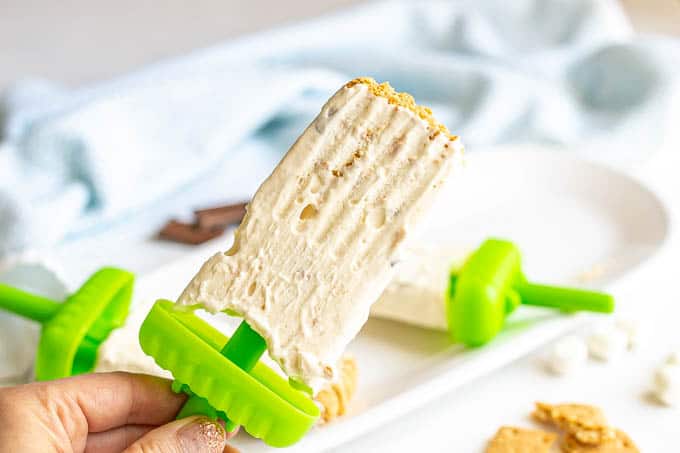 A hand holding a homemade s'mores popsicle with a green stick base and graham cracker crumbs on the top
