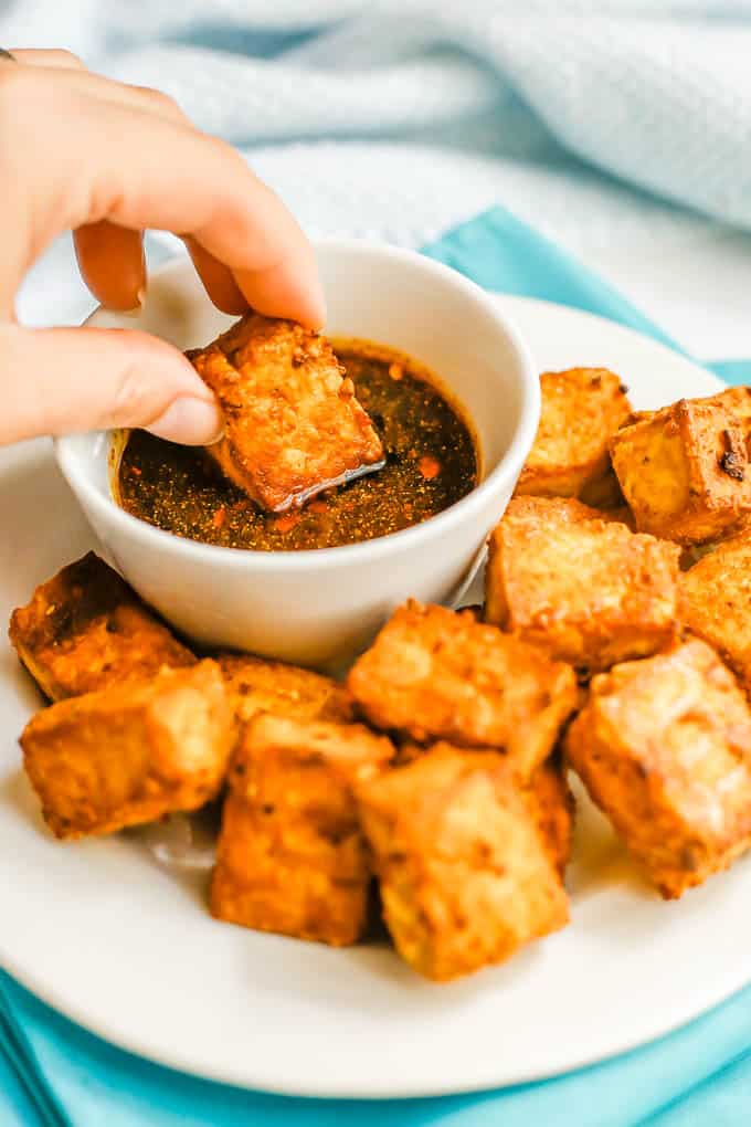 A hand dipping a crispy tofu piece into a soy sauce dipping sauce