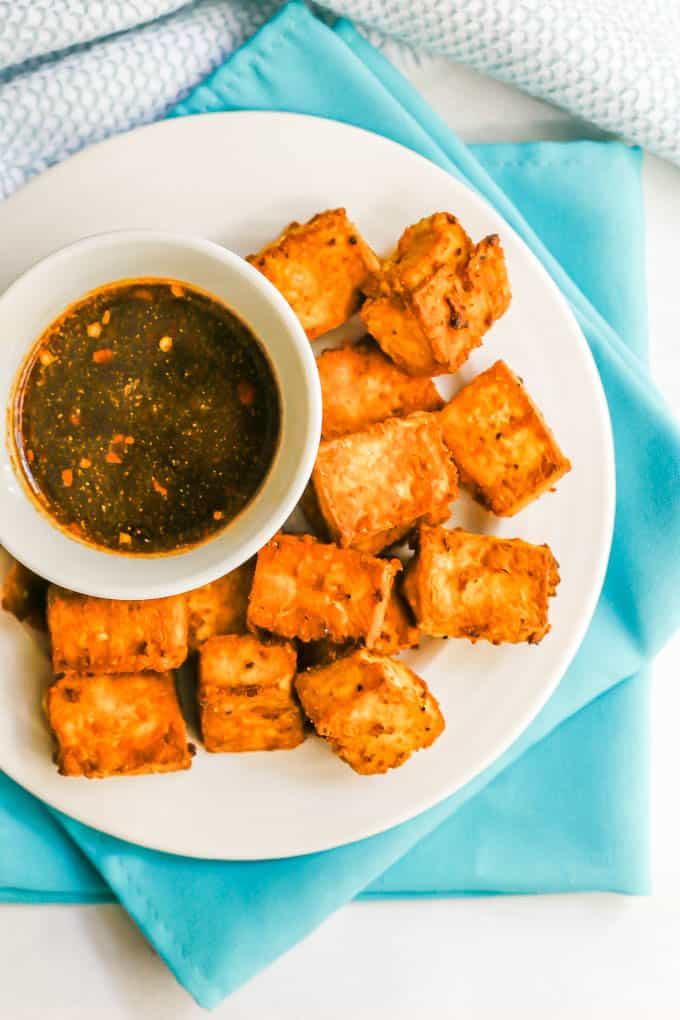 Close up of crispy tofu on a white plate with a small bowl of a brown soy sauce dipping sauce