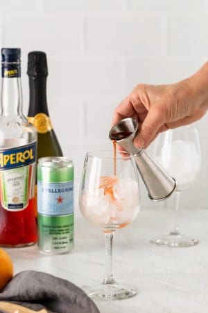 A shot glass pouring out Aperol into a wine glass with ice