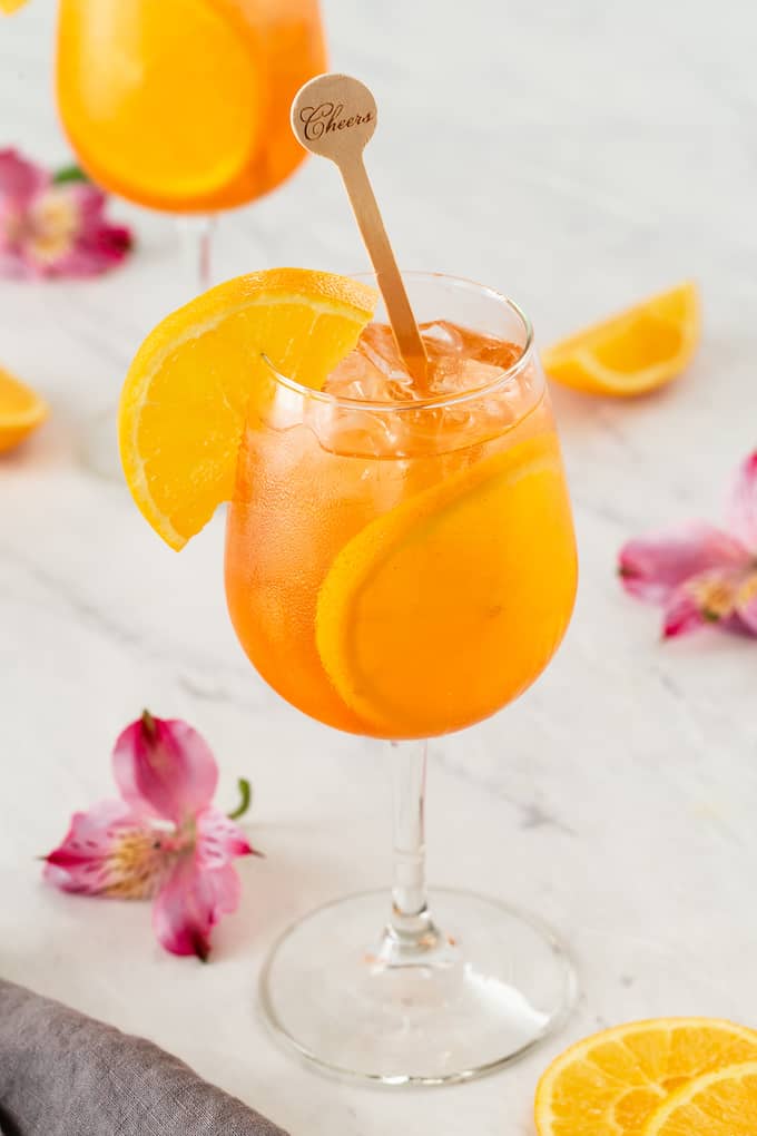 Two glasses of Aperol Spritz in wine glasses garnished with orange slices with pink flowers on the counter