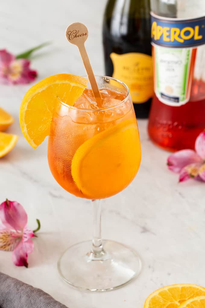 An Aperol Spritz cocktail with ice in a wine glass with orange slices and bottles in the background
