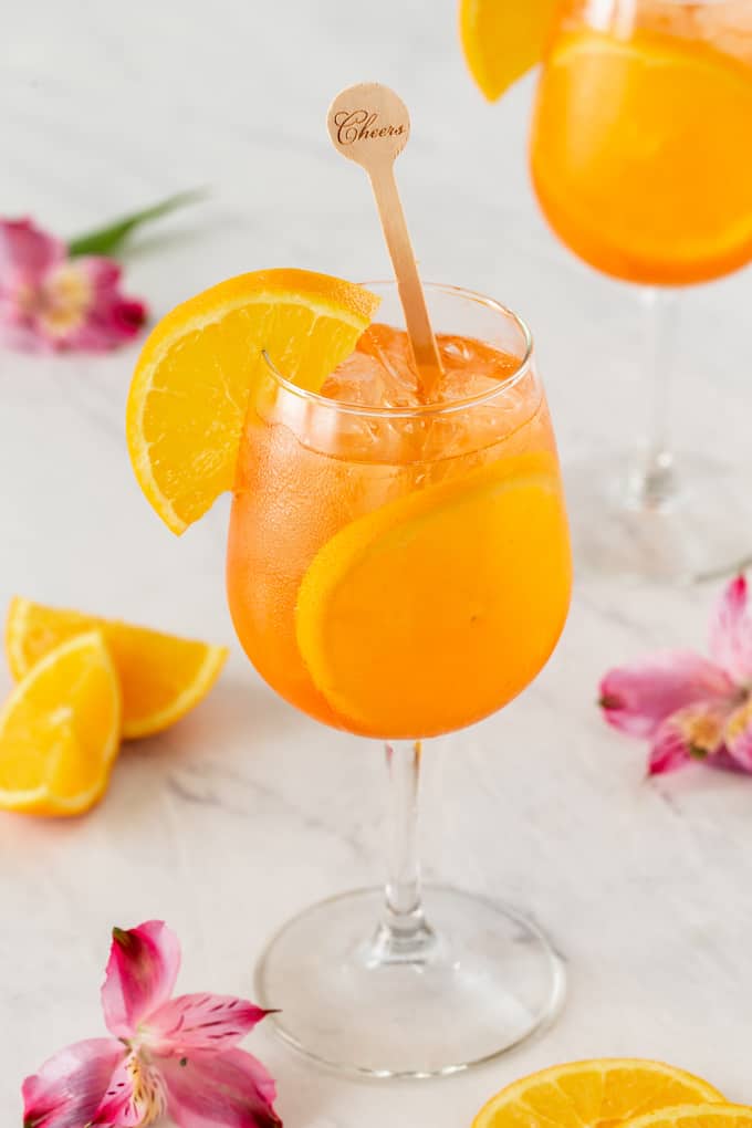 Two glasses with Aperol Spritz cocktail and orange slices and a wooden stir spoon that says Cheers on it