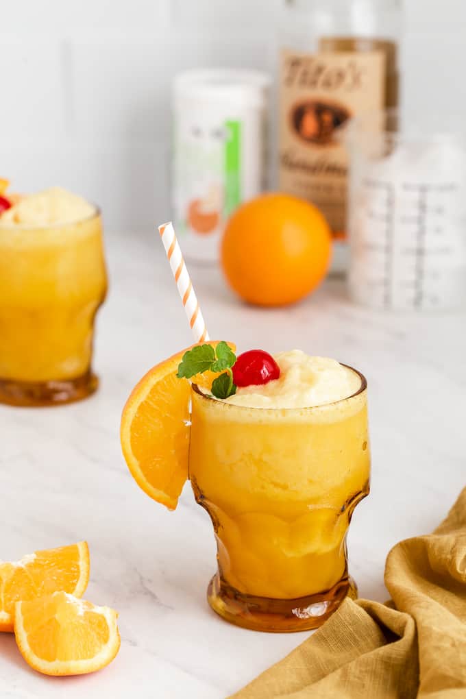 An alcoholic orange slushy in a glass with orange, cherry and mint garnishes and an orange striped straw, plus the drink ingredients in the background