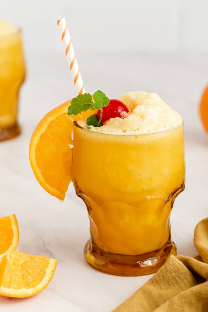 An amber glass with a slushy frozen screwdriver garnished with orange slice, cherry and a sprig of mint