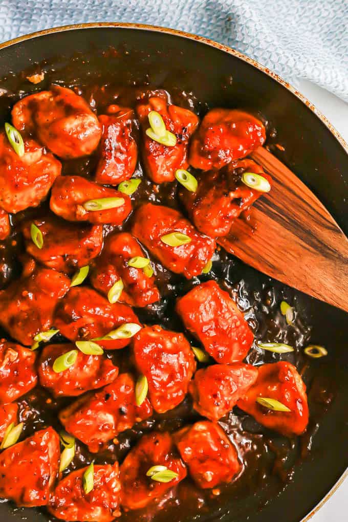 A wooden spatula scooping up some pieces of saucy orange chicken from a large skillet