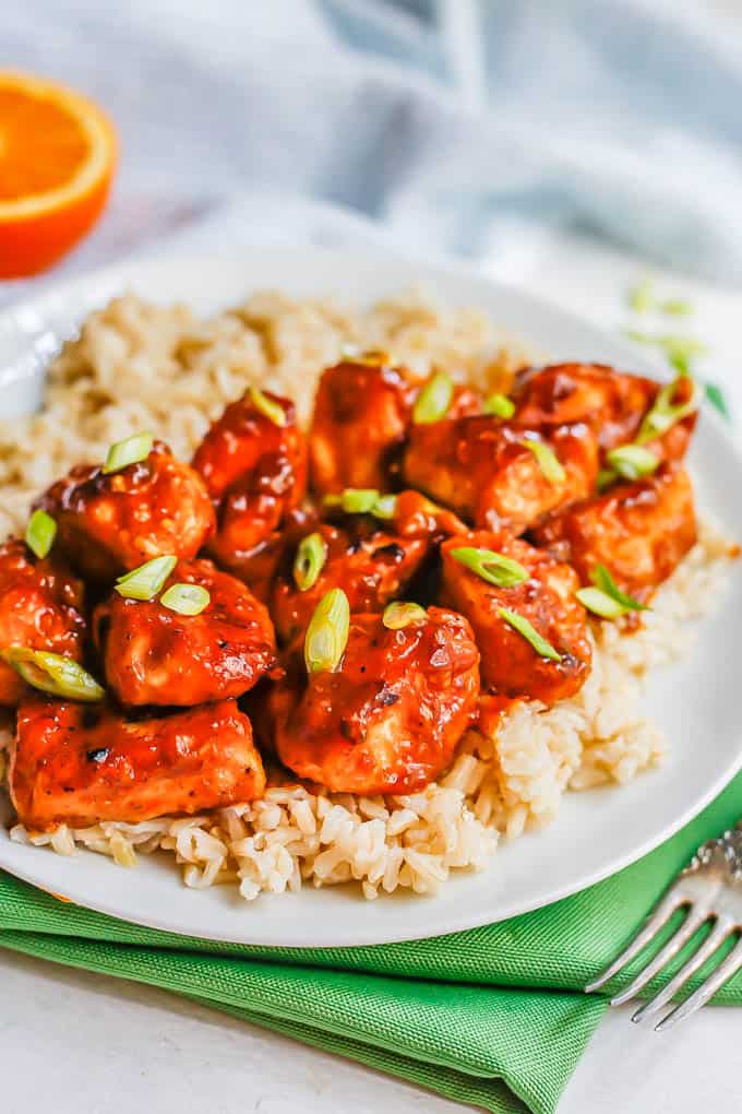 Close up of cubed orange chicken pieces served over steamed brown rice on a white plate with green onions sprinkled on top