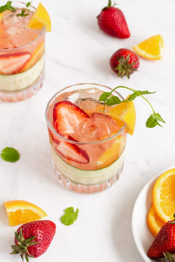 Two clear glasses with a cocktail mix and fresh fruit, plus fresh fruit nearby