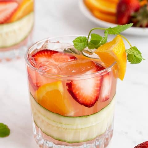 A short, clear drink glass with fruit, cucumber and Pimm's Cup cocktail with extra fruit laying around