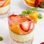 A short, clear drink glass with fruit, cucumber and Pimm's Cup cocktail with extra fruit laying around and a text overlay on the photo and another drink in the background.