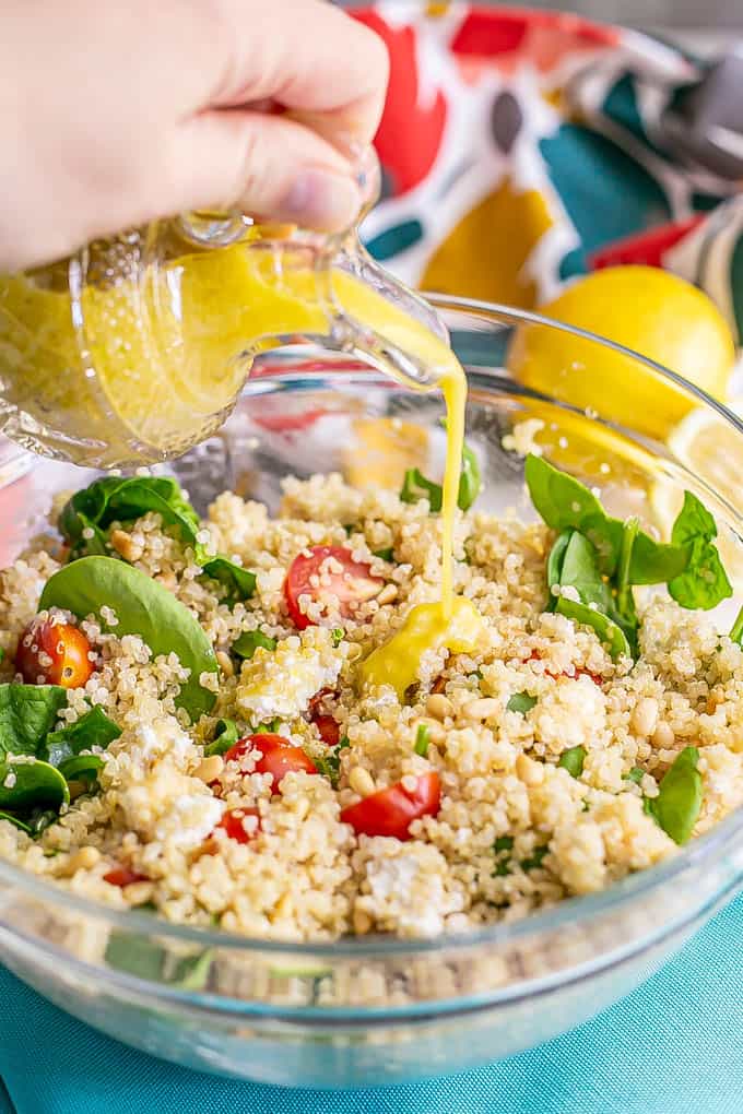 A hand pouring a lemony vinaigrette from a crystal jar over a quinoa salad with spinach and tomatoes