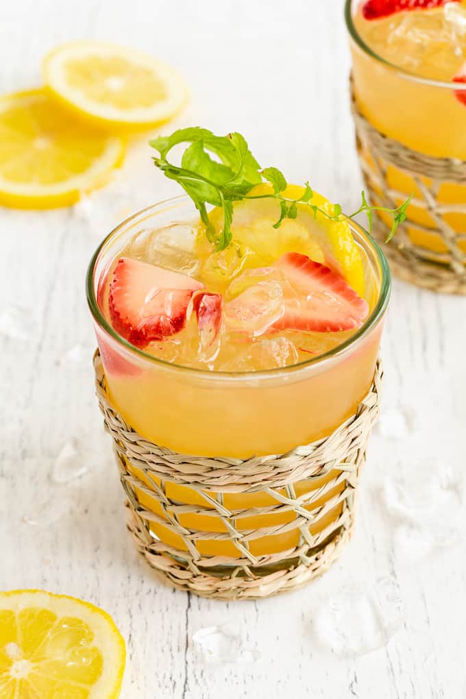A short clear glass with a lemonade tea and bourbon drink plus lemon and strawberry garnishes