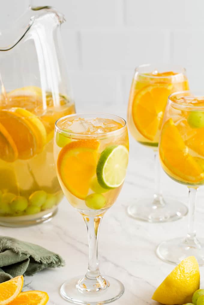 Three wine glasses filled with a fruity white sangria with orange and lime slices and grapes mixed in and a pitcher to the side