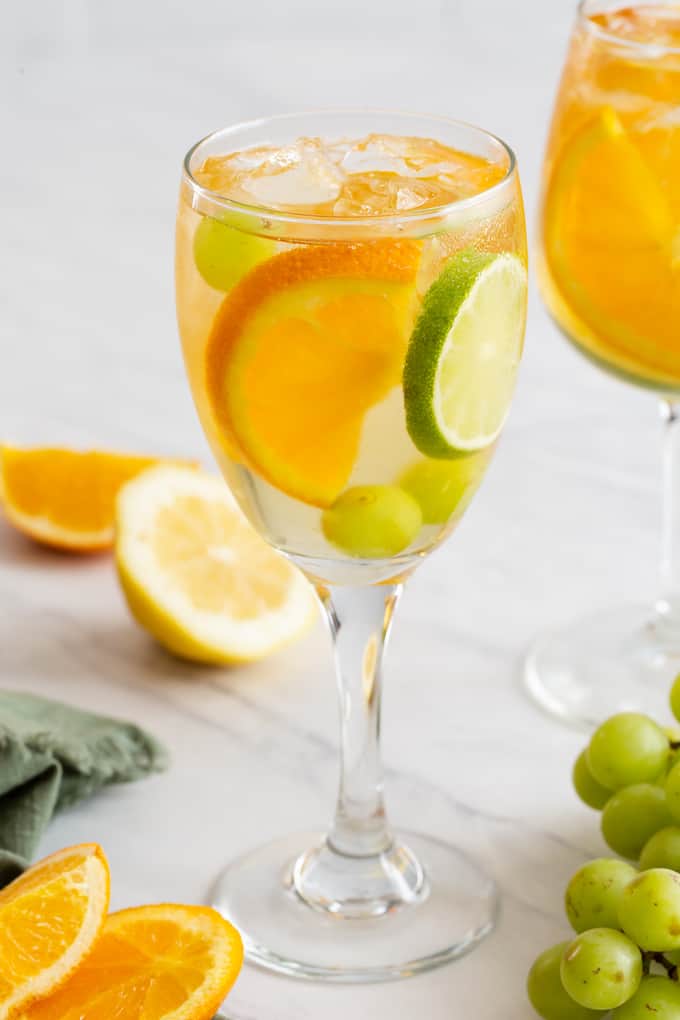 A wine glass filled with a white sangria, grapes, orange and lime slices and extra fruit lying nearby