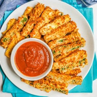 Overhead shot of crispy zucchini fries on a white plate with a small white bowl of marinara sauce