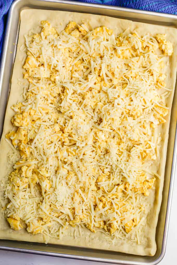 A sheet pan with a pizza dough spread out and topped with scrambled eggs and shredded cheese, before being baked