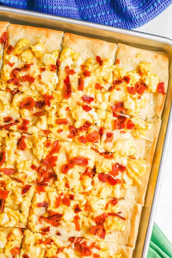 Overhead close up shot of a sheet pan with a cooked pizza with scrambled eggs, bacon and cheese, cut into slices