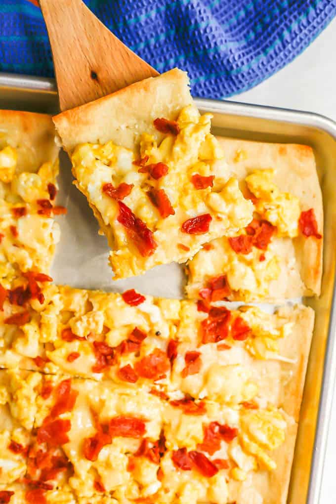 A spatula lifting up a piece of egg and bacon pizza from a sheet pan