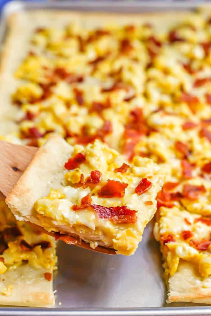 A wooden spatula lifting a slice of egg and bacon breakfast pizza from a sheet pan
