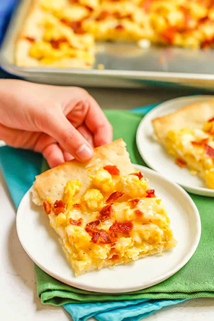 A hand picking up a piece of breakfast pizza from a small white plate