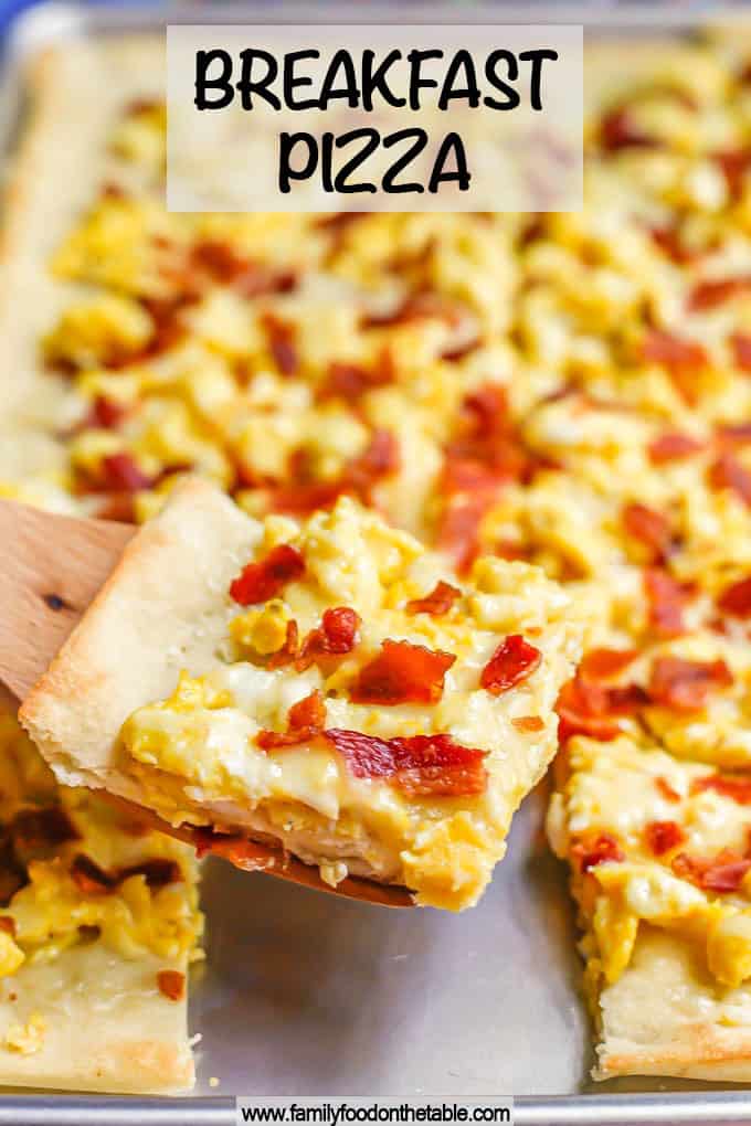 A wooden spatula lifting a slice of egg and bacon breakfast pizza from a sheet pan with a text overlay on the photo