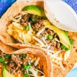 Egg and sausage breakfast tacos in a soft tortilla shell with cheese, avocado and green onion on top and a text overlay on the photo