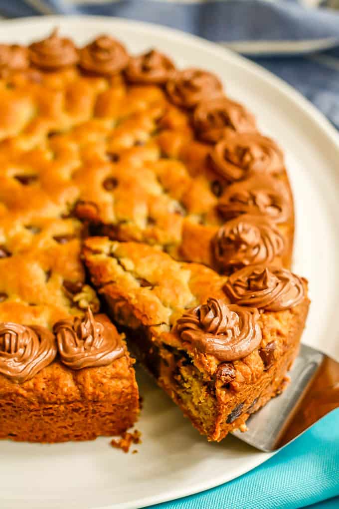 Close up of a slice of chocolate chip cookie cake with chocolate frosting being lifted from a white platter