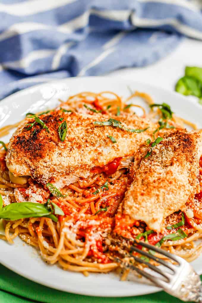 A sliced chicken Parmesan cutlet served over spaghetti noodles with basil sprinkled on top and a fork resting to the side of the plate