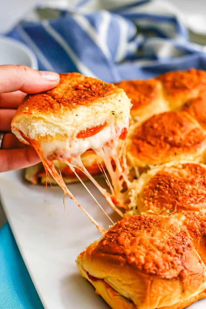A hand picking up a pizza slider with pepperoni and melty mozzarella cheese from a platter of sliders