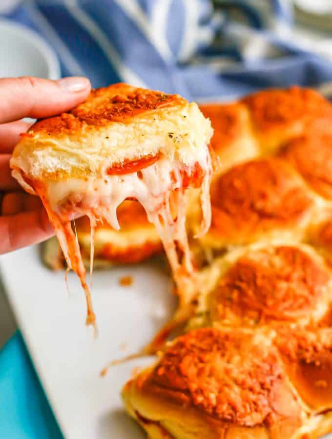 A hand holding up a cheesy pepperoni pizza slider from a plate of sliders