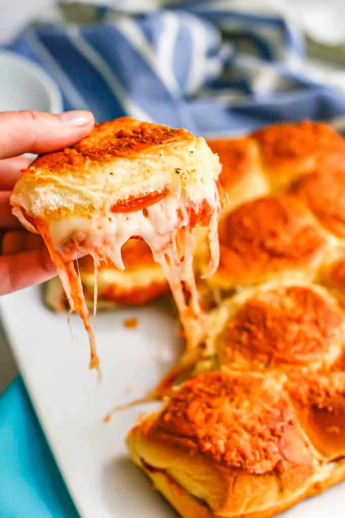 A hand holding up a cheesy pepperoni pizza slider from a plate of sliders