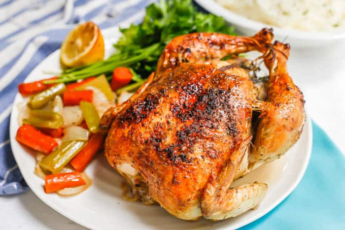 A whole roasted chicken served on a white platter with roasted veggies and lemon and parsley