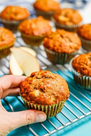 A hand about to pick up a whole grain apple cider muffin from a cooling rack