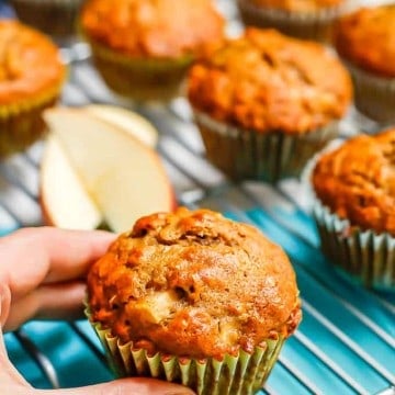 A hand about to pick up a whole grain apple cider muffin from a cooling rack