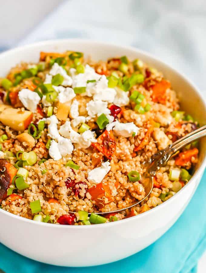 A large white bowl with a grain and veggie mix of quinoa, sweet potatoes, apples, feta cheese and green onions with a spoon resting in the bowl