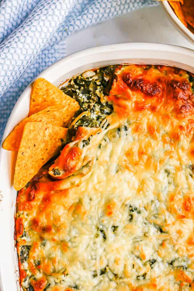 A browned and cheesy baked spinach dip in a white casserole dish with tortilla chips tucked into the corner for dipping