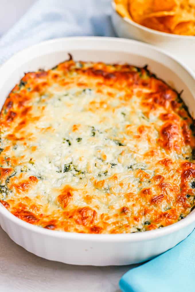 A white oval baking dish with browned, cheesy baked healthy spinach dip and a bowl of chips in the background