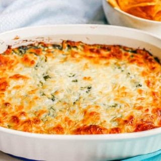 A white oval baking dish with browned, baked healthy spinach dip and a bowl of chips in the background