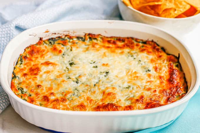 A white oval baking dish with browned, baked healthy spinach dip and a bowl of chips in the background